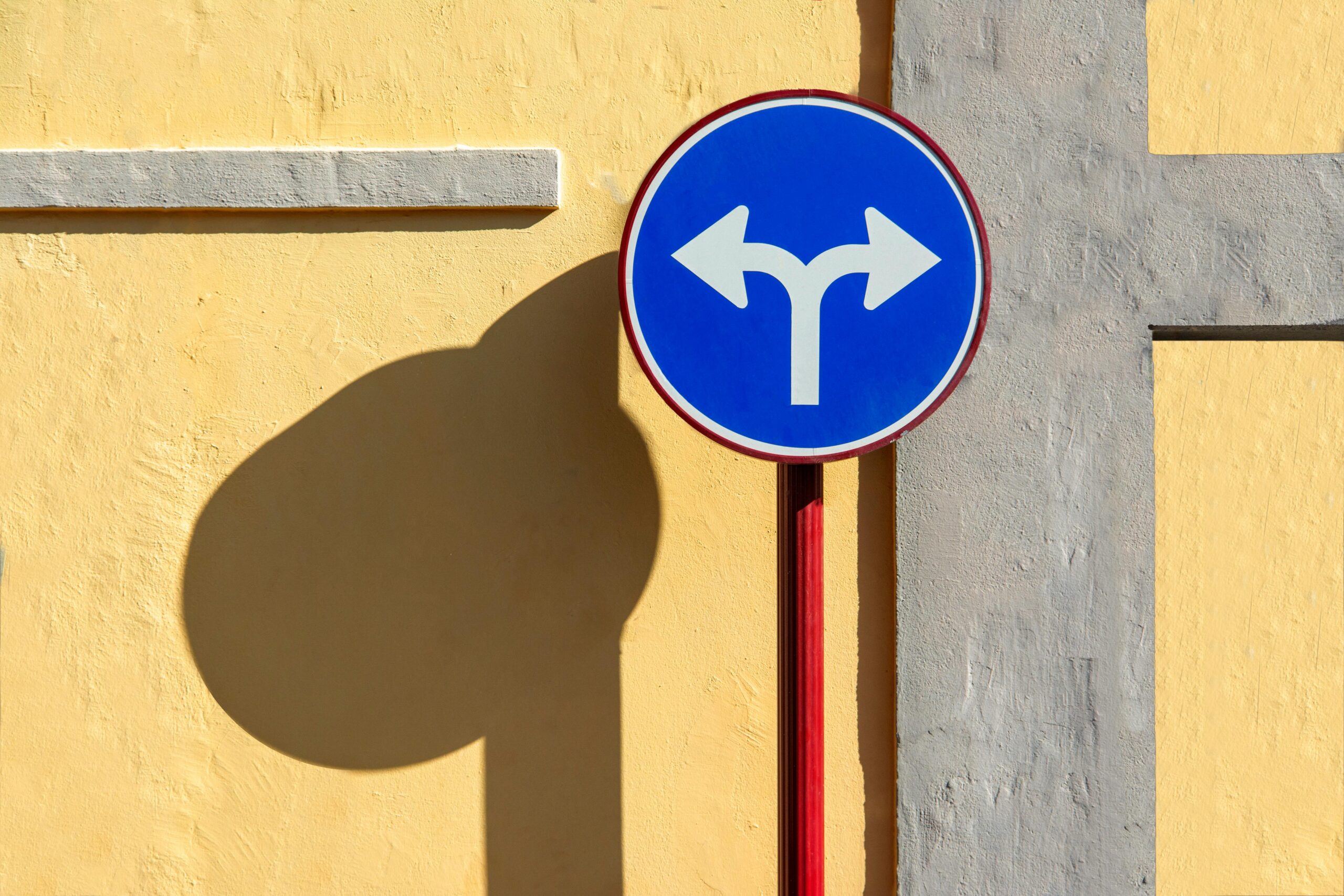 Blue traffic sign casting shadow on a vibrant yellow wall with strong sunlight.